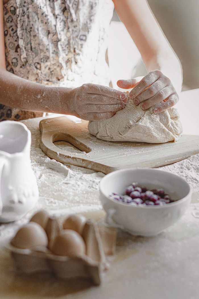A person kneads dough on a wooden board in a bright kitchen setting, surrounded by flour and kitchen items.