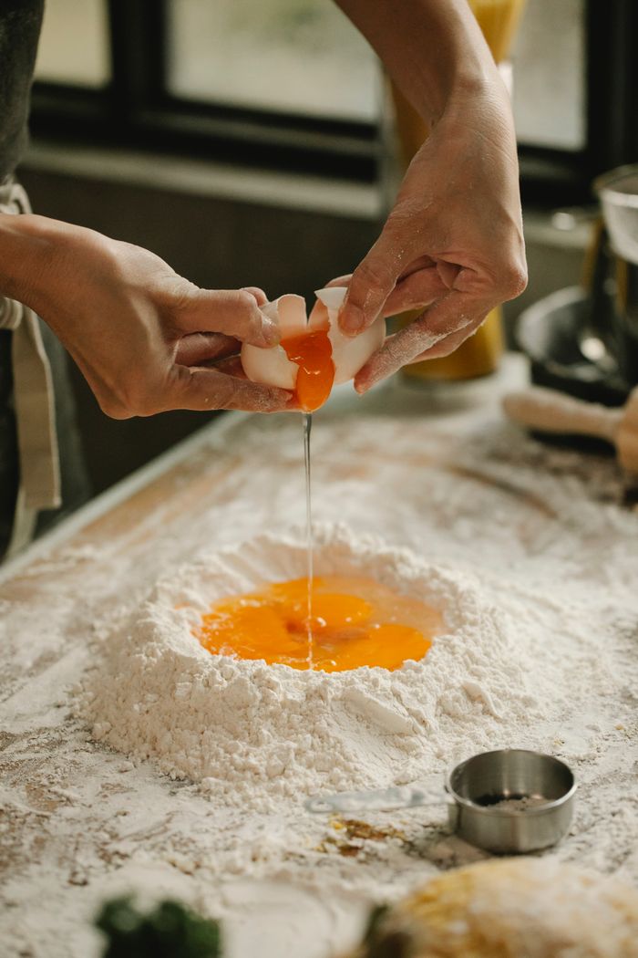 High angle of unrecognizable woman cracking egg into pile of flour making dough on table in kitchen