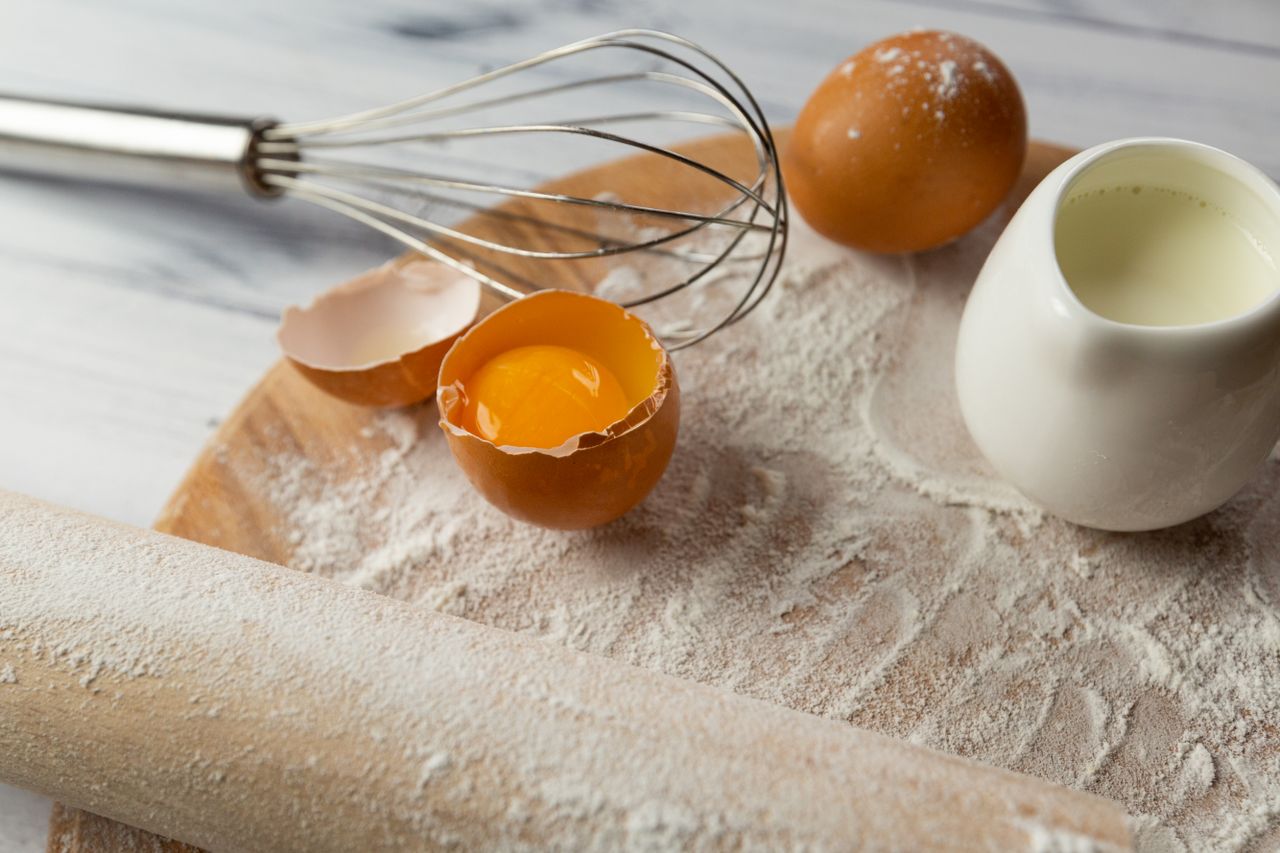 A close-up of baking essentials including eggs, flour, and a whisk on a wooden board.